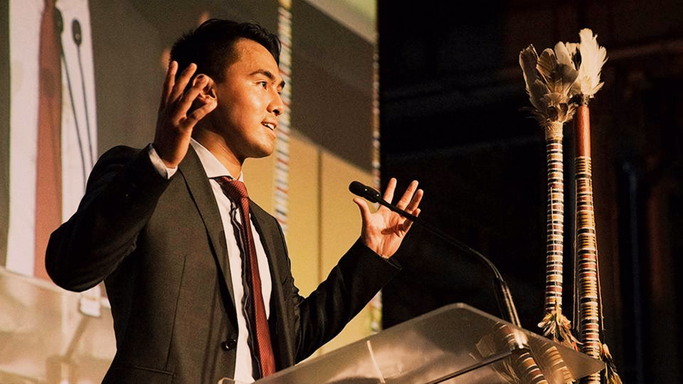 A CareerTrackers speaker gesturing while addressing an audience from a lectern, with First Nations ceremonial staffs beside him.