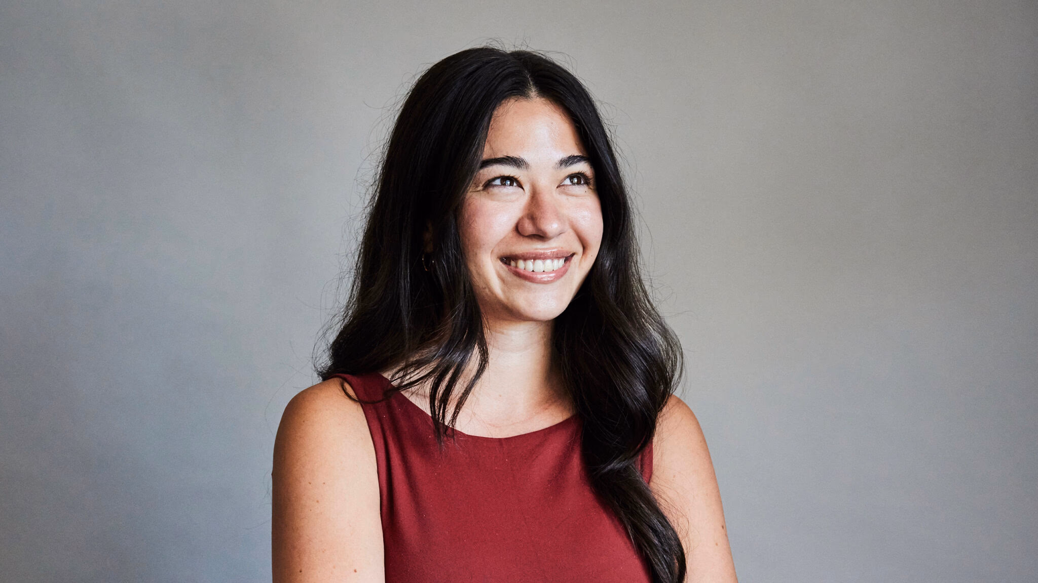 A smiling young woman with long dark hair looking off to the side against a plain studio backdrop.