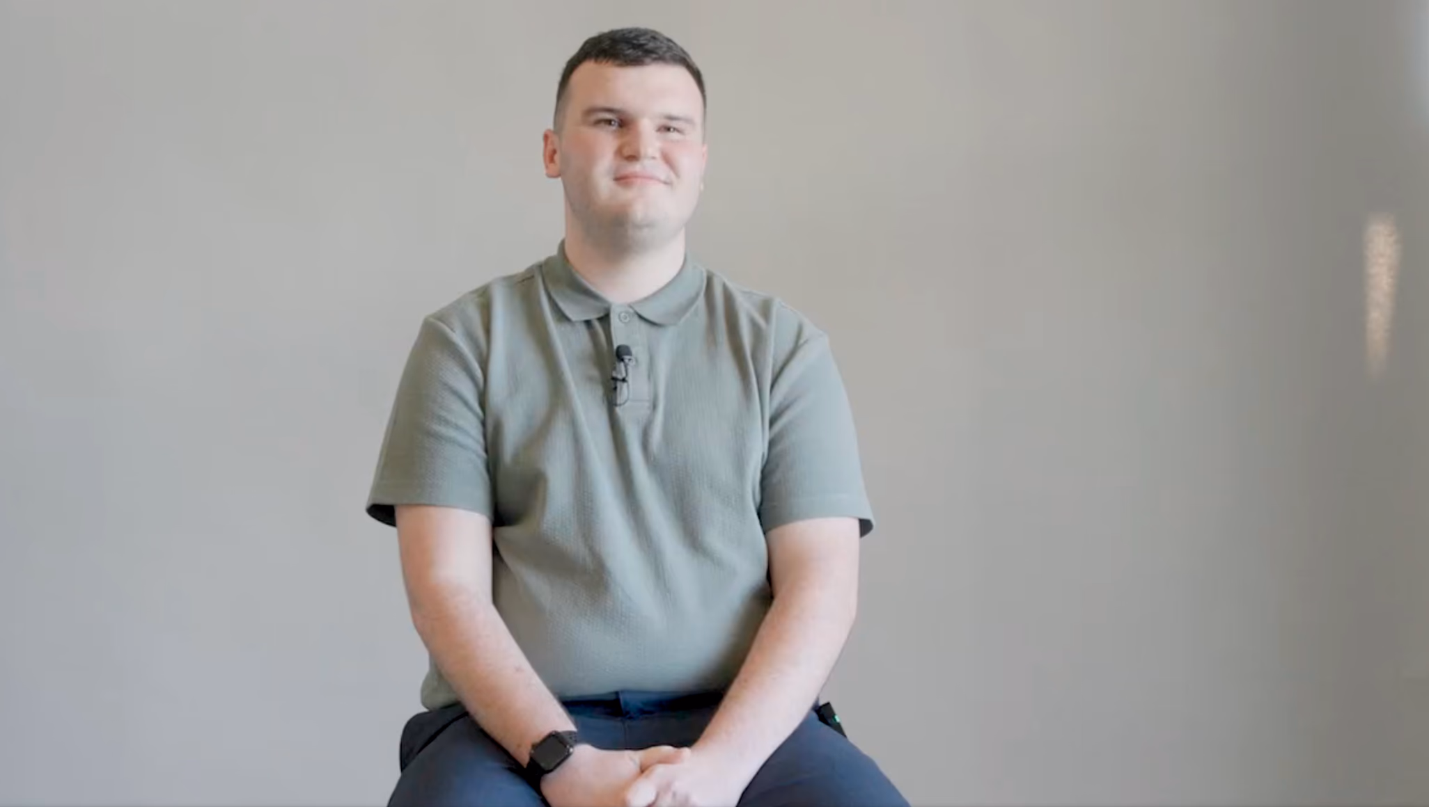 Still from a CareerTrackers student video — a young man in an olive polo shirt seated against a plain wall, smiling at the camera.