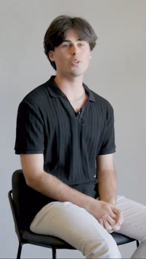 Still from a CareerTrackers reel — a young man in a black polo shirt seated against a plain wall, mid-sentence.